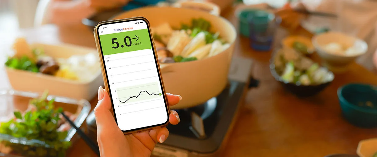 A hand holds a smartphone above a freshly prepared healthy dinner, the screen displaying a glucose reading
