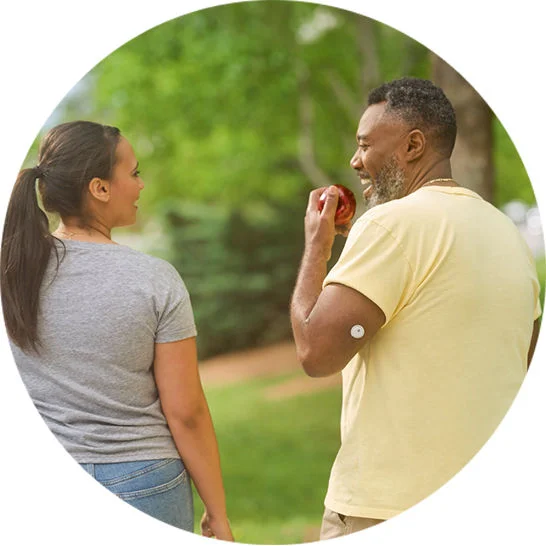 Two people walking outdoors, with one person eating an apple while wearing a continuous glucose monitor.