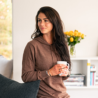 En person med langt hår og brun hættetrøje holder en hvid kop. Baggrunden viser en hvid hylde med bøger og en vase med gule blomster.