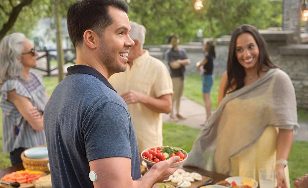 A man at an outdoor gathering holds a bowl of vegetables, with a FreeStyle Libre sensor on his upper arm.