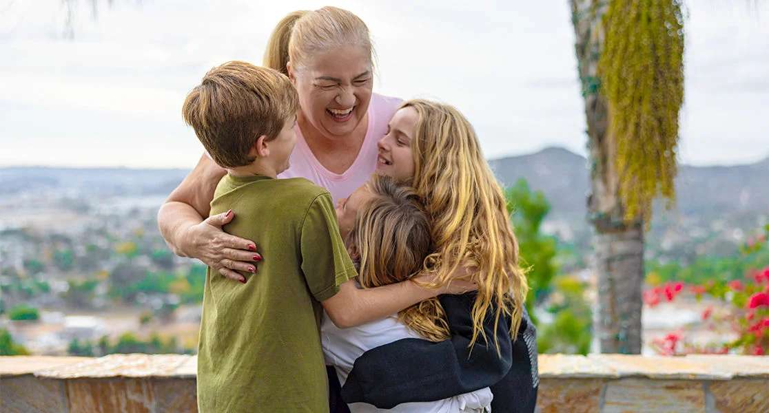 A woman and three children sharing a group hug outdoors with a scenic hillside view in the background.