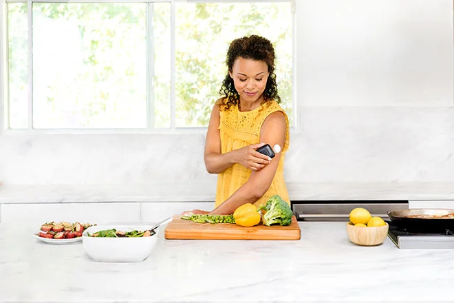 Mulher vestindo uma blusa amarela, em uma cozinha com vegetais, enquanto mede seus niveis de glicose por meio do leitor FreeStyle Libre