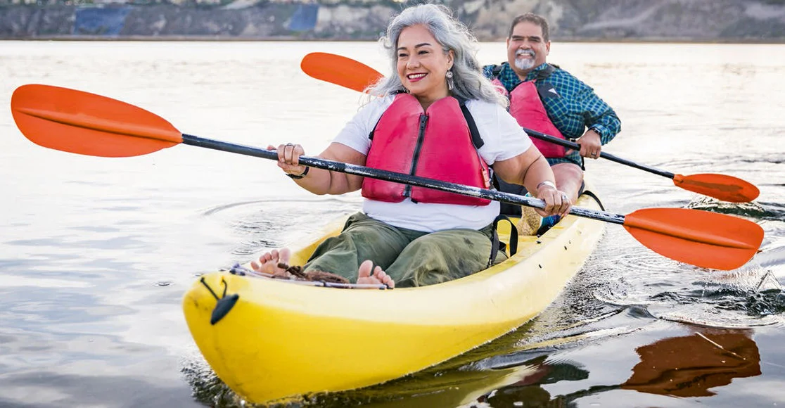 Two people wearing life vests paddle a yellow tandem kayak across calm water near a shoreline.