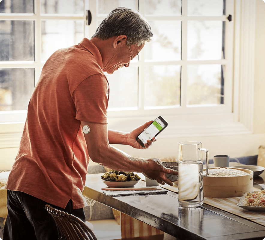一位老先生在廚房忙碌，右手臂配戴 FreeStyle Libre2 傳感器，左手拿著智慧型手機，視線看向手機畫面上 LibreLink 應用程式的葡萄糖數值。