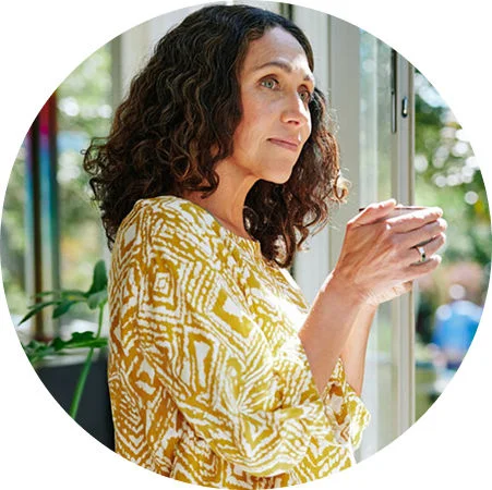 A woman holding a cup while standing near a sunlit window with indoor plants in the background.