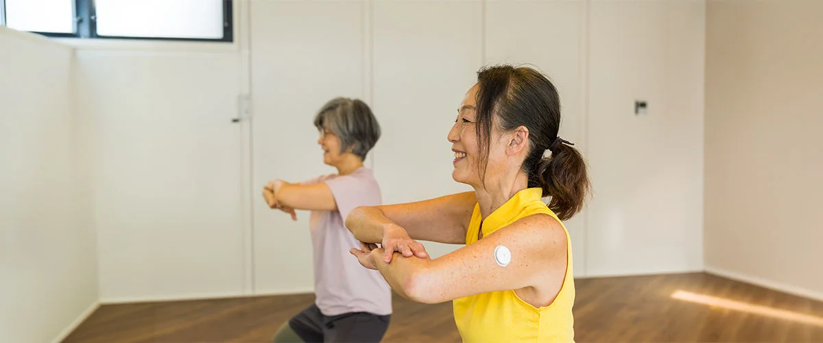 Hai người tập vận động trong phòng, một người đeo cảm biến đường huyết trên cánh tay, minh họa theo dõi HbA1c.