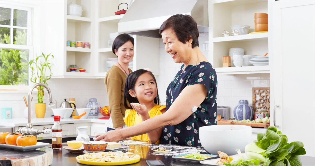 A grandmother cooks with her granddaughter in a bright kitchen, with a FreeStyle Libre sensor on her upper arm.