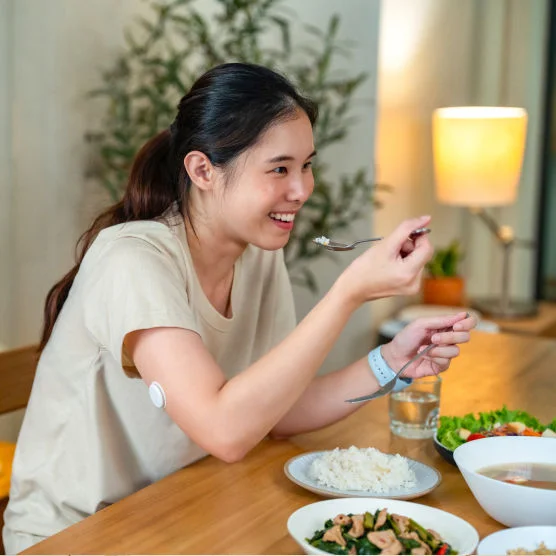 Mulher sentada à mesa durante uma refeição, levantando uma colher para comer, com o sensor Freestyle Libre no braço.