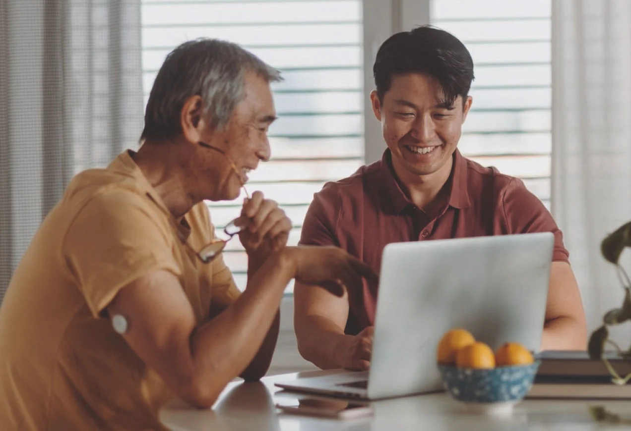 Persona utilizando una computadora mientras otra señala, con un sensor de glucosa visible en el brazo.