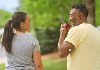Un hombre y una mujer caminan al aire libre mientras él come una manzana, con un sensor FreeStyle Libre visible en la parte superior de su brazo.