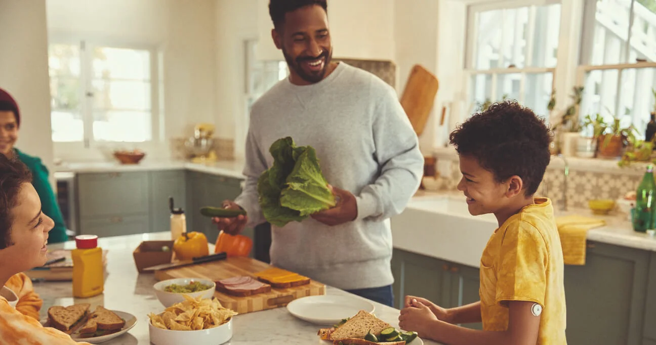 Familia reunida en la cocina durante la merienda; el padre cocina mientras su hijo, que usa el sensor FreeStyle Libre, lo ayuda con alegría