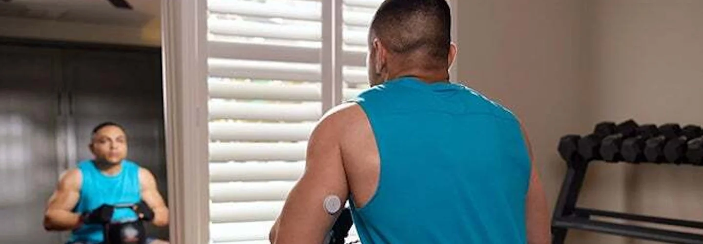 Homem usando uma camiseta azul e realizando um exercicio fisico com pesos em uma sala. Em seu braço há um sensor FreeStyle Libre