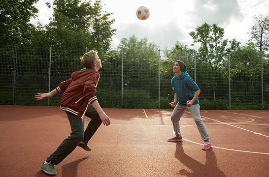 Two people playing with a ball on an outdoor sports court surrounded by trees and fencing.
