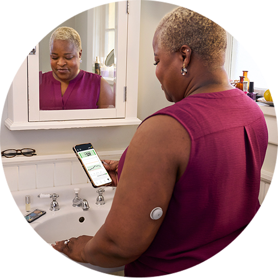 Person viewing glucose data on a smartphone in a bathroom, wearing a CGM sensor on the upper arm.