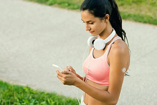  Mulher com fones de ouvidos apoiados no pescoço, verificando seu smartphone após realizar uma atividade física. Ela usa um sensor FreeStyle Libre em seu braço
