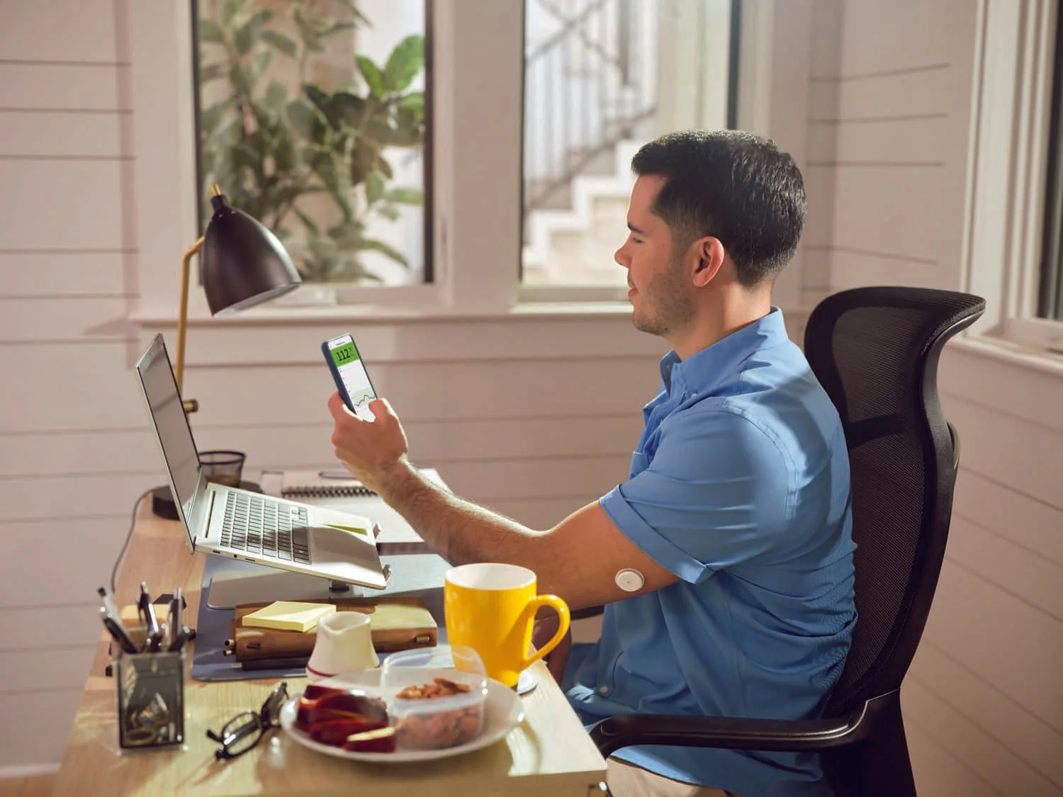 Rapaz mexendo no celular, sentado à mesa com café da manhã e notebook
