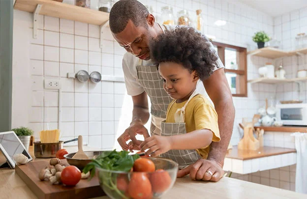 Um adulto e uma criança preparam uma refeição saudável na cozinha com legumes frescos sobre a bancada.