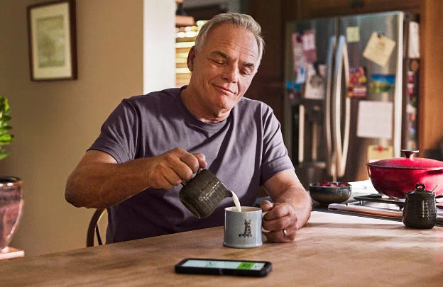 An elderly man adds milk to his coffee with a smartphone displaying the Libre app on the table beside him.