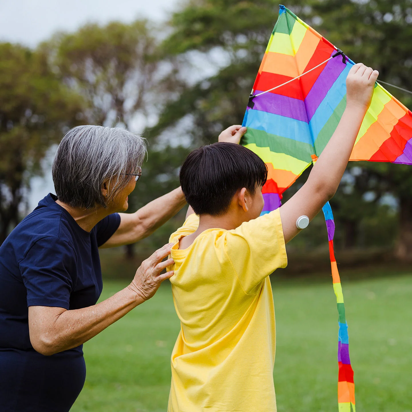 A boy wearing a FreeStyle Libre sensor on the upper arm while flying a colorful kite outdoors with another person in a park.