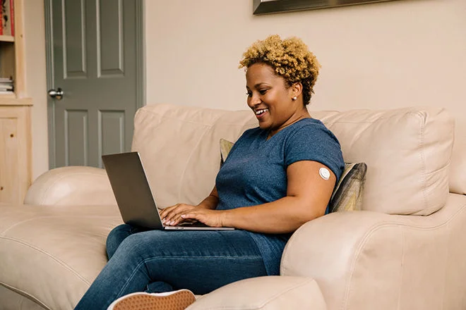 Mulher vestindo uma camiseta azul, sentanda em um sofá com o notebook em seu colo fazendo uma pesquisa. Ela usa um sensor FreeStyle Libre em seu braço