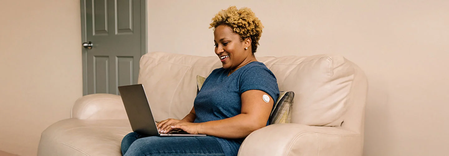 Mulher vestindo uma camiseta azul, sentanda em um sofá com o notebook em seu colo fazendo uma pesquisa. Ela usa um sensor FreeStyle Libre em seu braço
