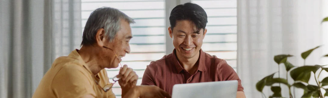 Duas pessoas sentadas em uma mesa olhando para um laptop, em um ambiente interno iluminado com plantas ao fundo.