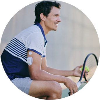 Person seated on a tennis court holding a racket, wearing a continuous glucose monitor on the upper arm.