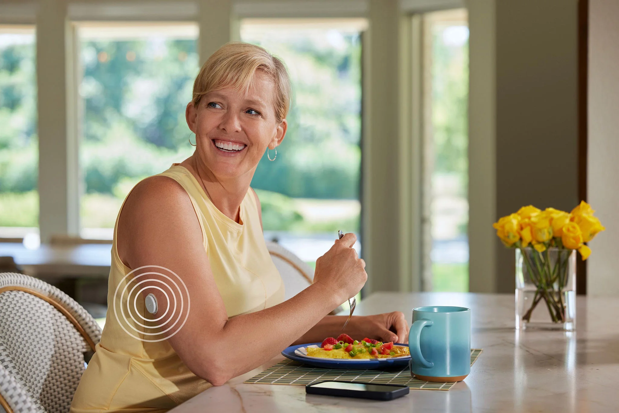 Mulher vestindo blusa amarela sentada à mesa para comer. Ela segura um garfo e tem diante de si um prato com uma omelete, uma caneca e um celular. Há um sensor FreeStyle Libre em seu braço direito