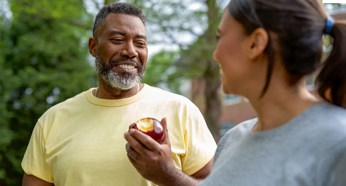 Two people talking outdoors in a park-like setting, with one person holding a partially eaten red apple.