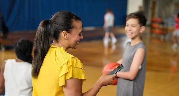 Person holding a smartphone while talking with a child holding a basketball in a gym, illustrating LibreLinkUp glucose data sharing.