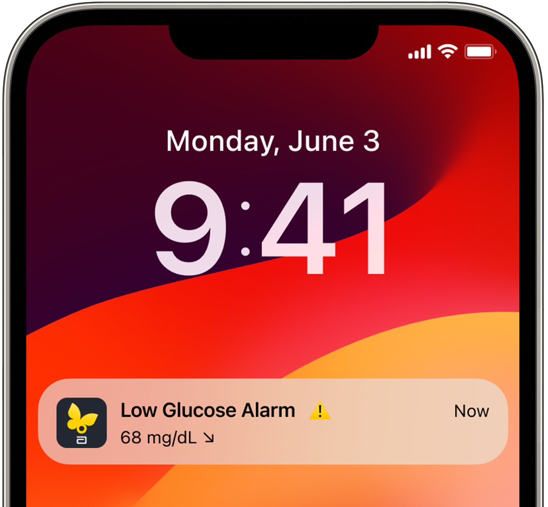Smartphone on a nightstand displaying a low glucose alert notification beside a juice box, glasses, and stacked books in a dimly lit bedroom.