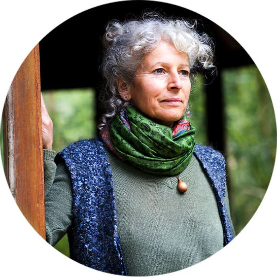 A woman standing in a doorway outdoors, wearing a green scarf and blue vest with trees in the background.