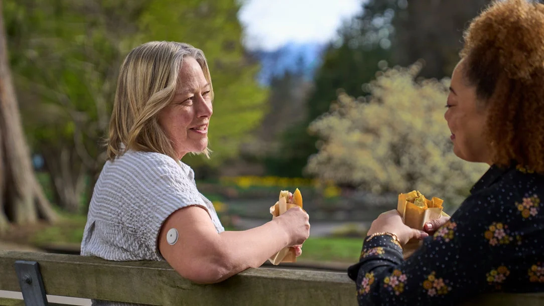 Mujer con el sensor FreeStyle Libre conversando con una amiga al aire libre mientras comparten un aperitivo saludable.
