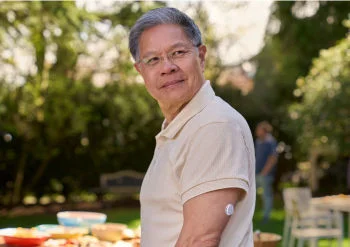 一名中年男士站在戶外庭院，左手臂佩戴 FreeStyle Libre2 傳感器，自信地看向遠方，背後的桌子上放滿食物。