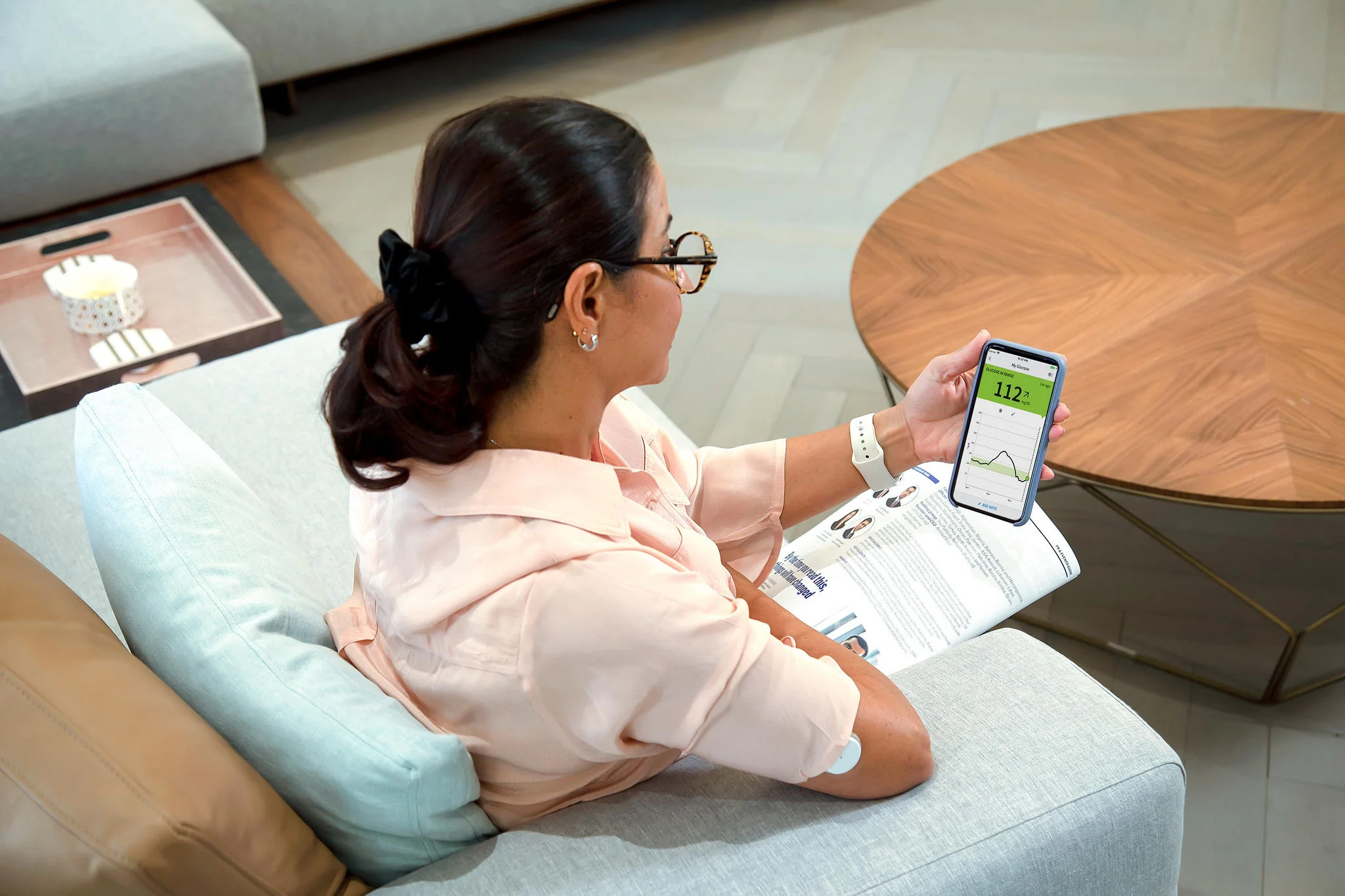 Mulher sentada em seu sofá de cor azul clara, vestindo uma camisa na cor salmão, usando um relógio de pulseira branca e óculos de grau. Ela tem uma revista aberta em seu colo. Em sua mão, ela segura um smartphone com o aplicativo FreeStyle LibreLink e usa o sensor Freestyle Libre em seu braço direito