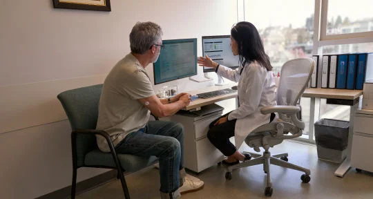 一名男性患者坐在診間內，與身穿白袍的醫師討論，兩人面對電腦螢幕，醫師手指向螢幕解釋 LibreView 上的數據。