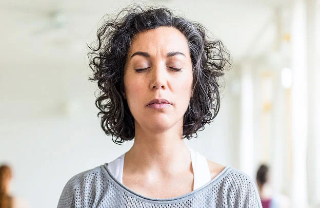 A woman with her eyes closed meditates while in a communal yoga class, a relaxing method of boosting holistic health.