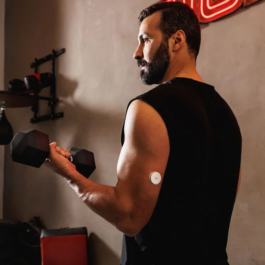 A man having a dumbbell in his hand, wearing a black shirt and a Freestyle Libre sensor on the back of his upper arm in the gym
