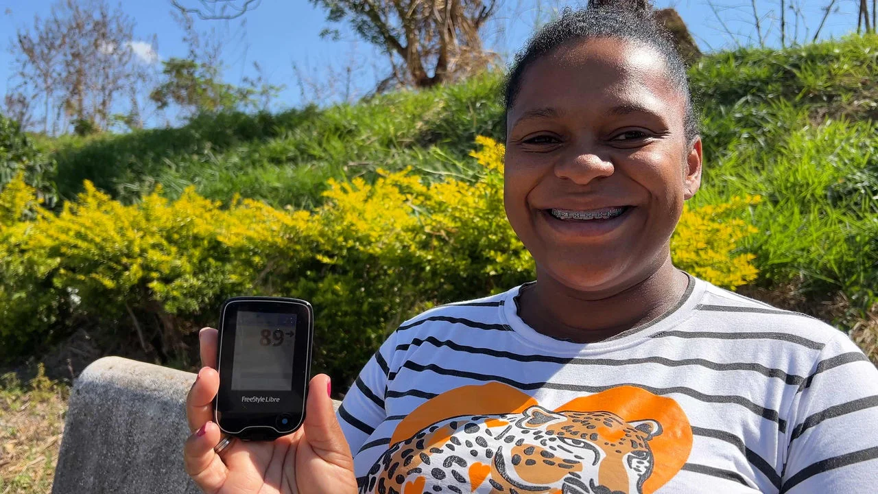 Mulher vestindo uma camiseta branca com listras pretas e uma ilustração de onça. Sentada em um banco de concreto em um parque, ela sorri e mostra o leitor FreeStyle Libre em sua mão