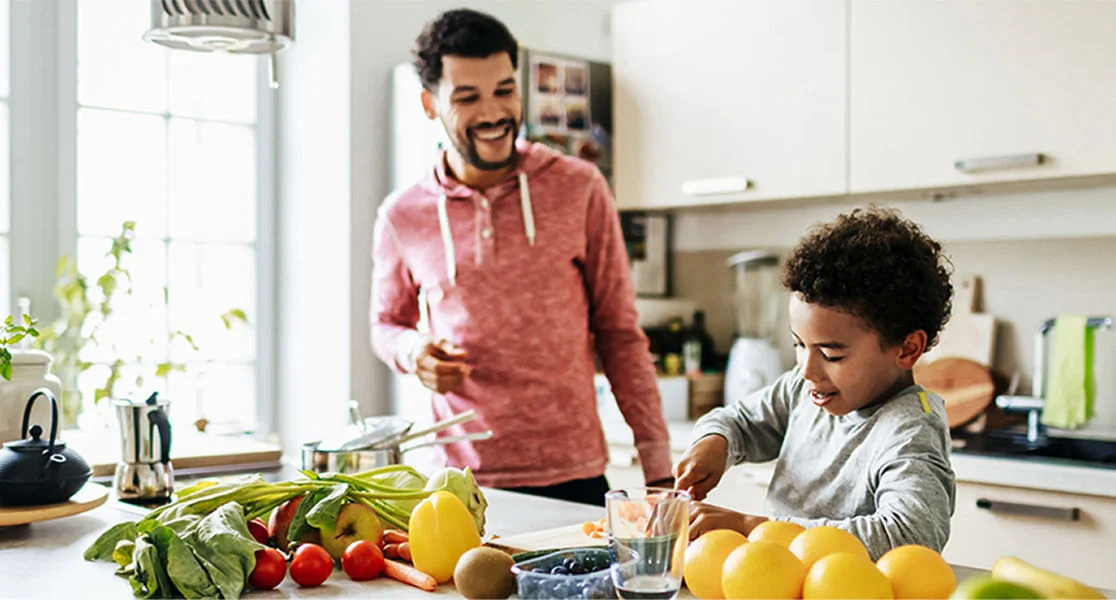 Person and child preparing fresh fruit together at a kitchen counter with oranges, berries, and vegetables.