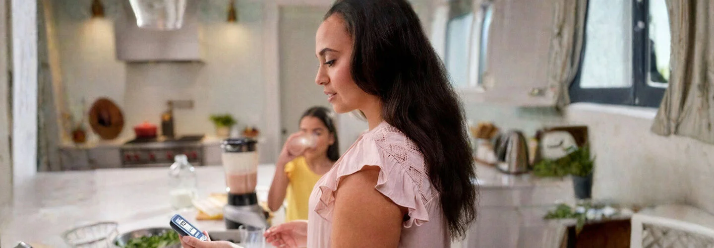 mulher em uma cozinha moderna e bem iluminada, segurando um leitor verificando seus níveis de glicose no aplicativo FreeStyle LibreLink em seu smartphone. Ela está próxima a uma bancada com folhas de espinafre em uma tigela, um liquidificador contendo líquido rosa, e outros utensílios. Ao fundo, outra pessoa está bebendo um copo, sugerindo um momento de rotina saudável.