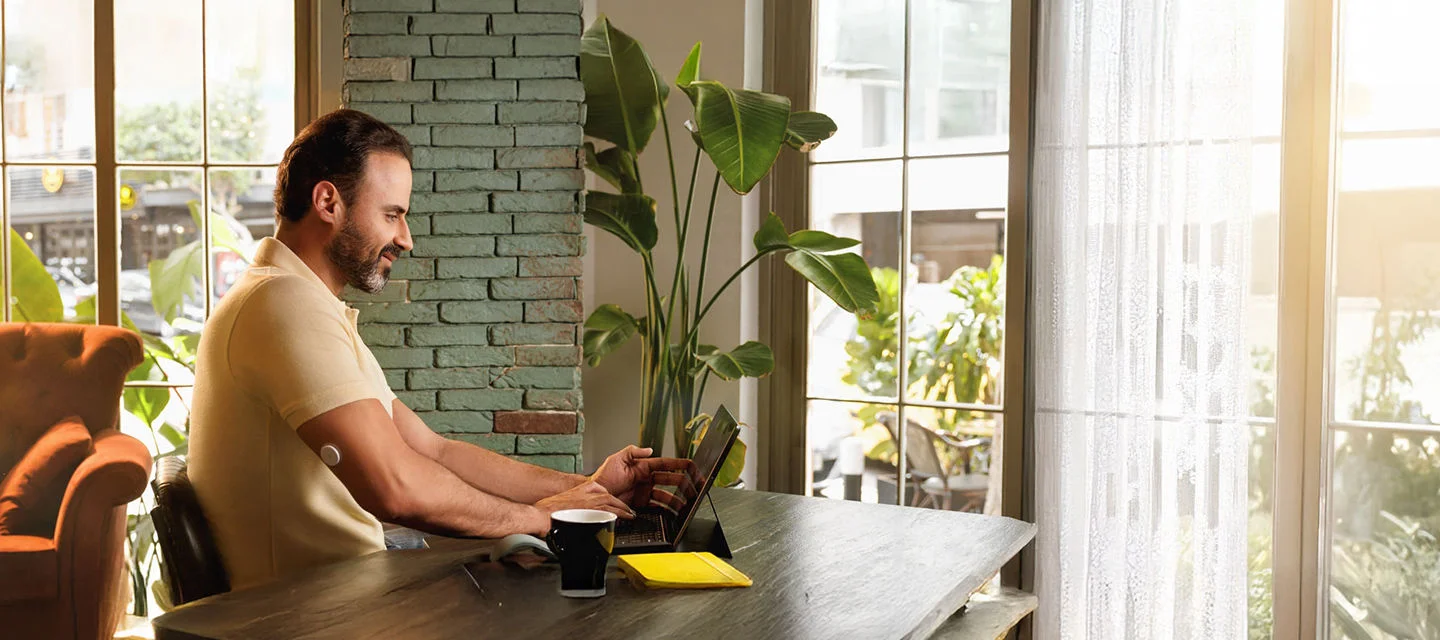 A man wearing a FreeStyle Libre sensor while sitting on a table using his laptop.