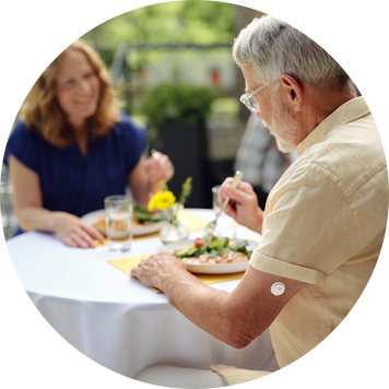 Two people seated at an outdoor table eating a meal, with plates of food and flowers in the background.