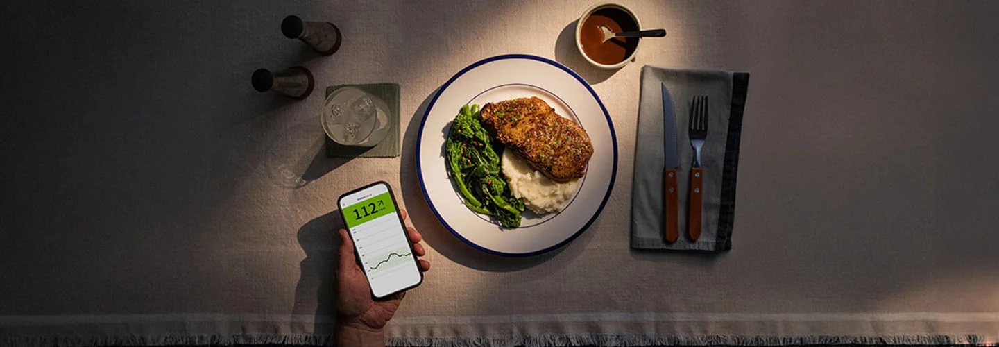 Mesa de refeição com proteína e vegetais e uma mão segurando o celular