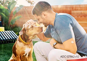 Homem ajoelhado ao ar livre acariciando um cachorro, com o sensor FreeStyle Libre visível no braço