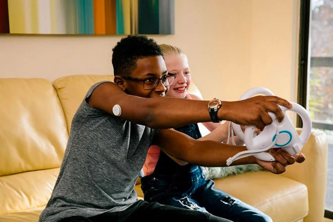 Dois jovens jogando video game em uma sala de estar, em um dos meninos há um sensor FreeStyle Libre em seu braço