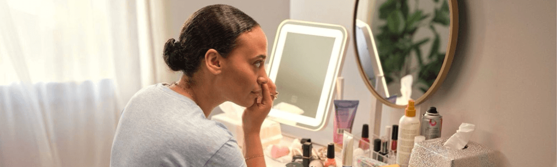 Mulher sentada à frente de uma penteadeira com diversos itens de cuidados pessoais, aplicando maquiagem. O ambiente é bem iluminado, com luz natural.