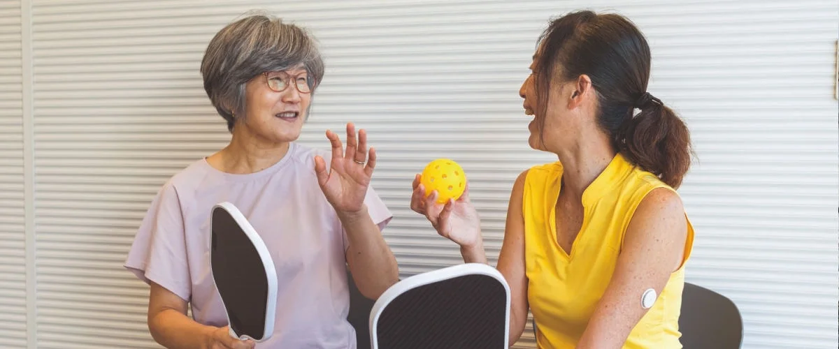 A Woman wearing a FreeStyle Libre sensor on the upper arm while participating in an indoor activity with another woman.