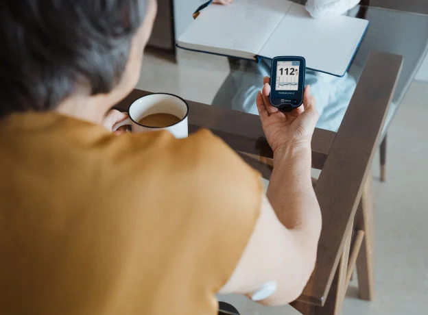 Persona sentada en una mesa sosteniendo un lector FreeStyle Libre 2 que muestra un nivel de glucosa, mientras toma café y tiene un cuaderno abierto al frente.