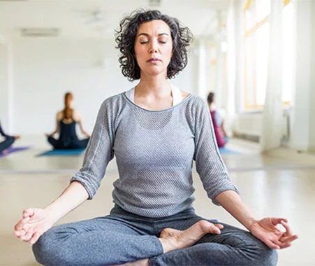 A woman seated cross-legged in a yoga studio during a meditation session, with others in the background.